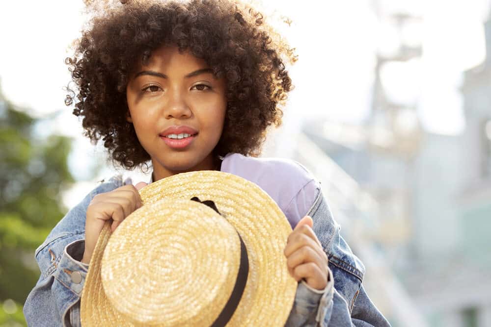 An attractive curly woman with a straw hat in her hands smiling toward the camera.