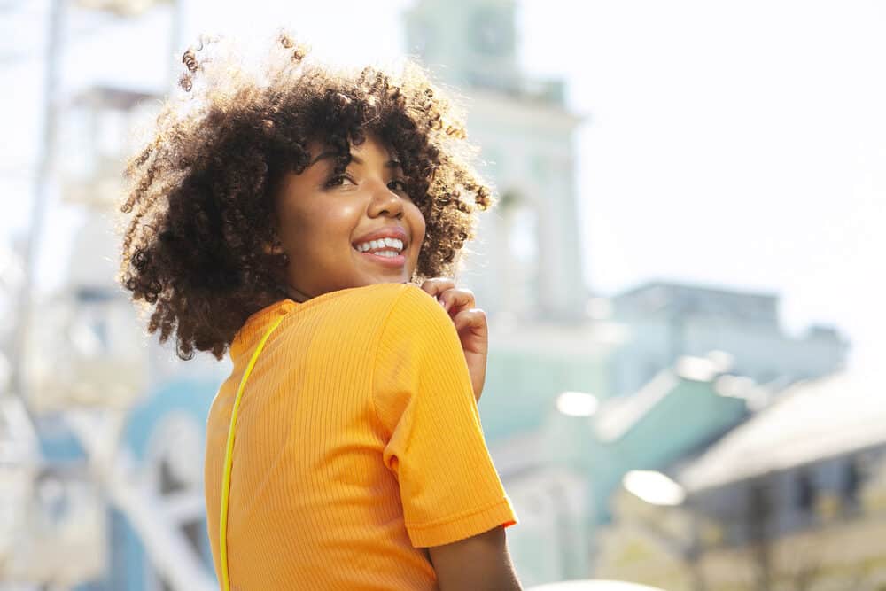 Happy black girl that's a hair care enthusiast with curly hair follicles wearing an orange shirt.