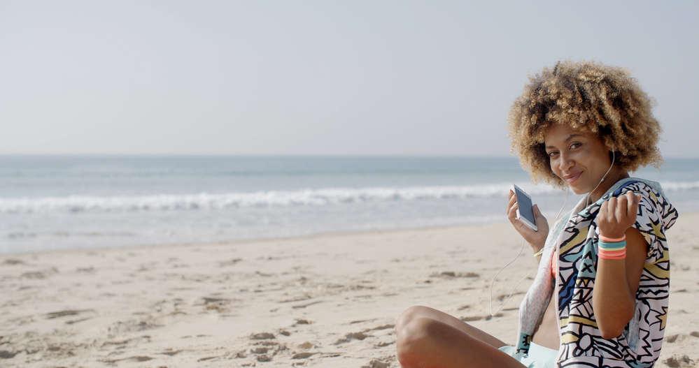 Light skin woman wearing a curly afro moisturized with coconut oil sitting on the beach listening to Apple headphones.