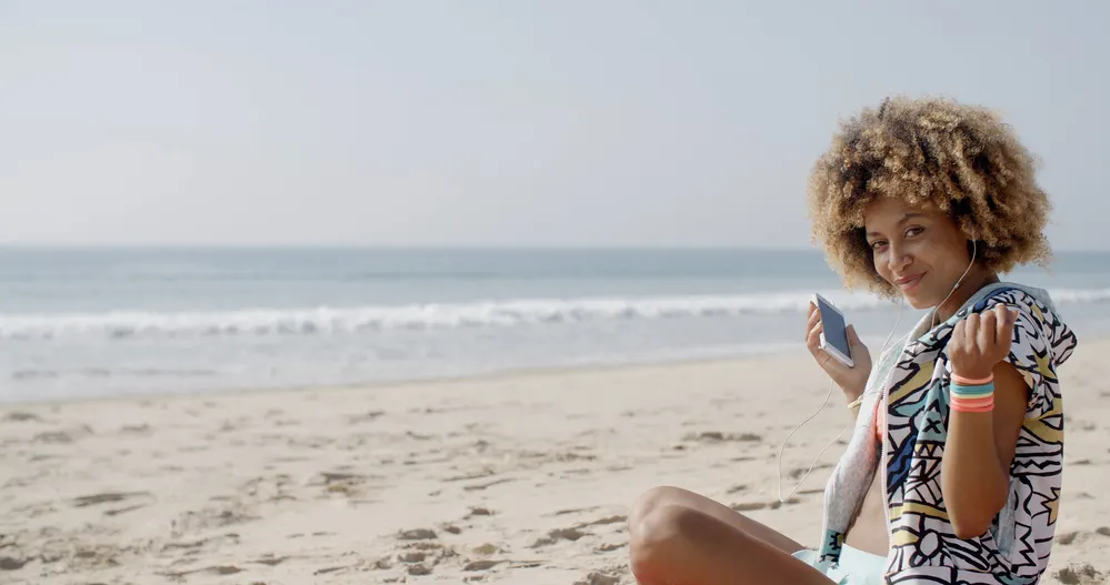 Will My Hair Fall Out If I Bleach It? Twice? Too Much? 3 Light skin woman wearing a curly afro moisturized with coconut oil sitting on the beach listening to Apple headphones.