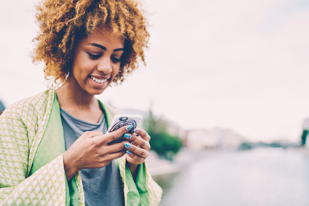 Cute black female with dark brown bleached hair colored with Splat dye using a mobile device outside on a fall day.