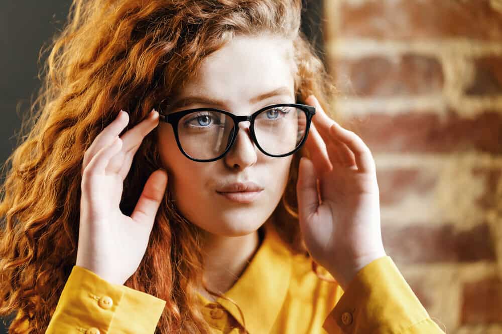 Curly redhead standing near a bricked wall wearing wavy bleached hair colored with Overtone.