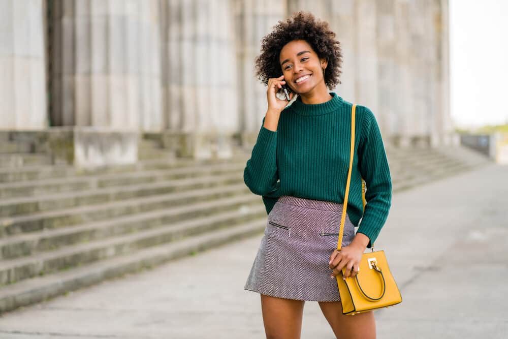 Black lady with a kinky, coily afro hairstyle wearing a green shirt, gray skirt, and yellow purse.