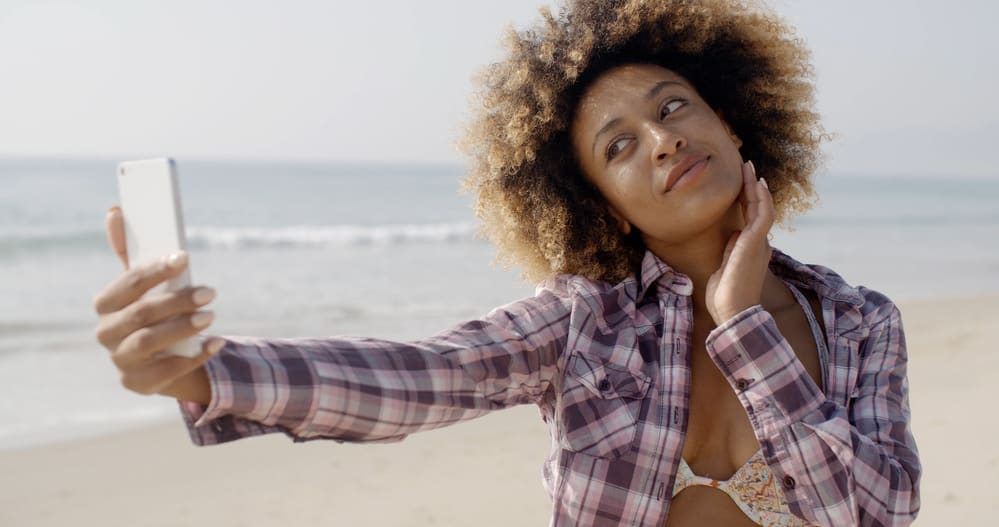 Black lady at the beach with damp hair taking a selfie photo with her new iPhone.