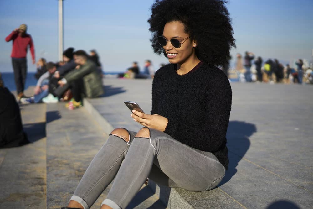African American female in gray jeans with a huge afro reading about the biotin hair growth supplement on her phone.
