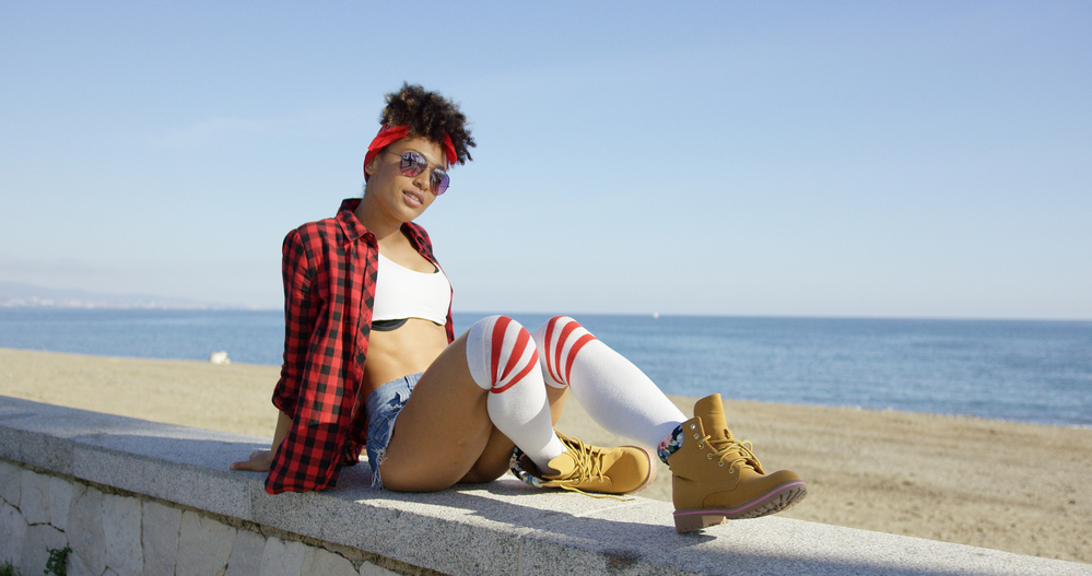 Trendy Black woman with damp hair relaxing on a beachfront wearing timberland boots.