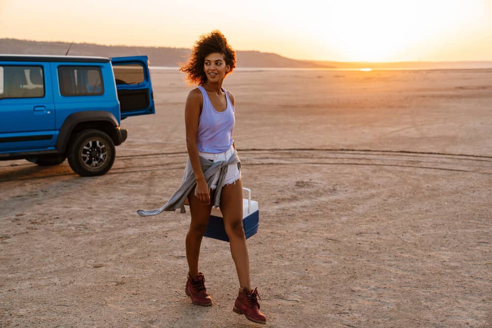 Black girl with curly red hair walking with a blue cooler outside in the sand.
