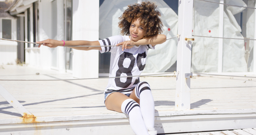 A black lady with bouncy brown and black 4C curls wearing an athletic white t-shirt and knee-high socks.