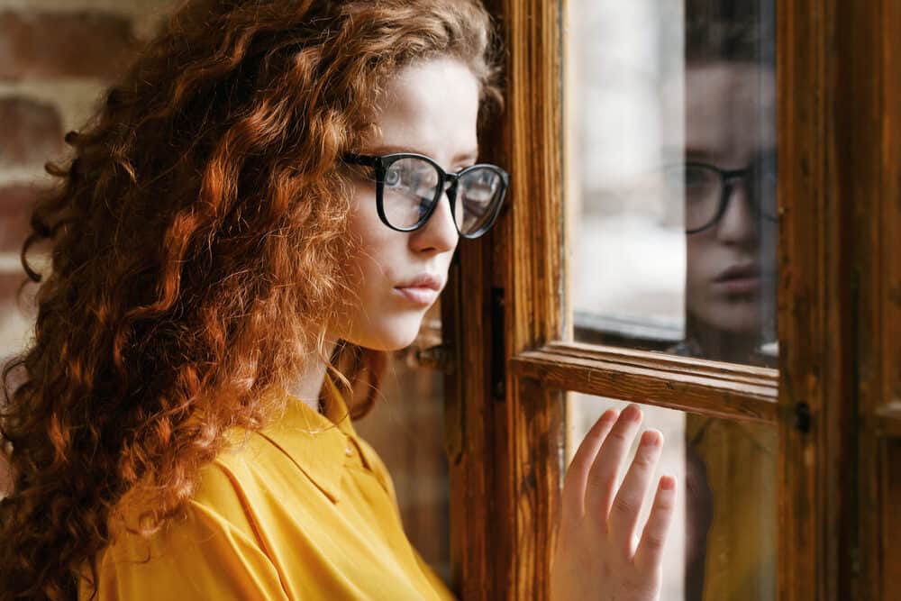 Attractive white girl with balayage-colored hair looking out of a wooden window seal.