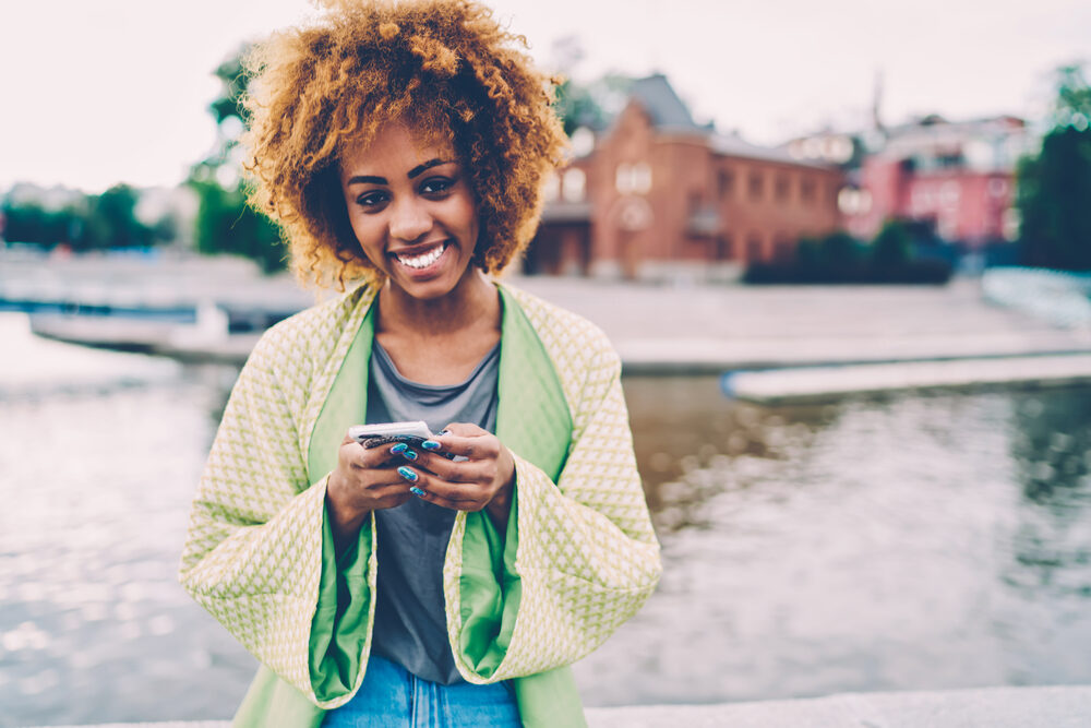 Cheerful woman with curly dark hair styled with permanent Splat hair dye standing near a lake.