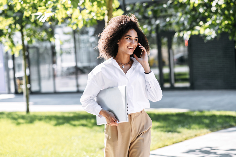 Cute black woman with a curly hair type talking on the phone while walking outdoors.