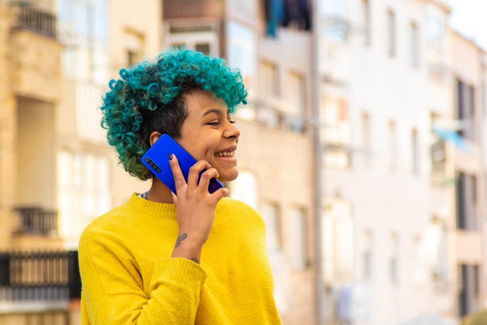 Young black girl with blue hair standing outside talking to a friend on the phone wearing a sweater.