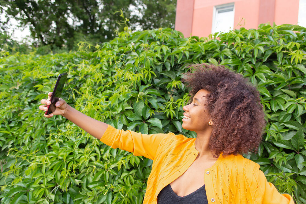 A young black woman takes a selfie outdoors after using a deep conditioning hair mask.