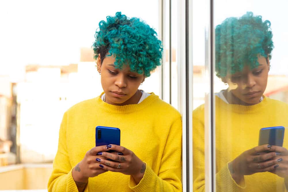 Black girl with naturally brown hair that has dyed her hair blue wearing a yellow sweater and purple fingernail polish.