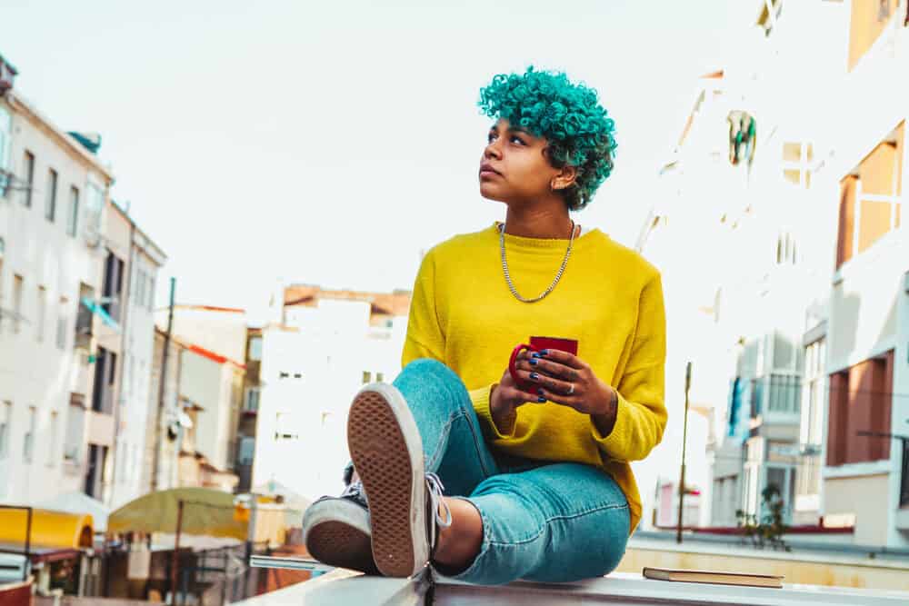 A modern young black lady with a blue hair color getting some sun exposure while drinking a cup of coffee.