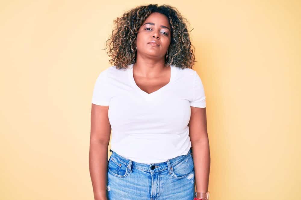 A black woman with subtle highlights wearing a gold watch, red bracelet, white t-shirt, and blue jeans.