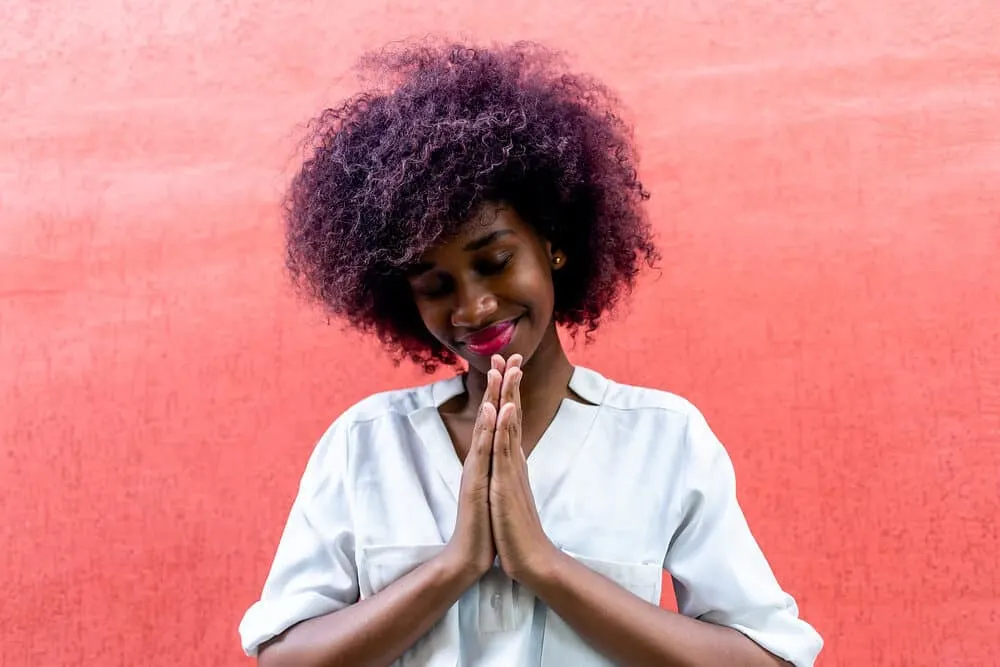 Black female making a gesture of gratitude with purple color-treated hair covering her dark brown natural color.