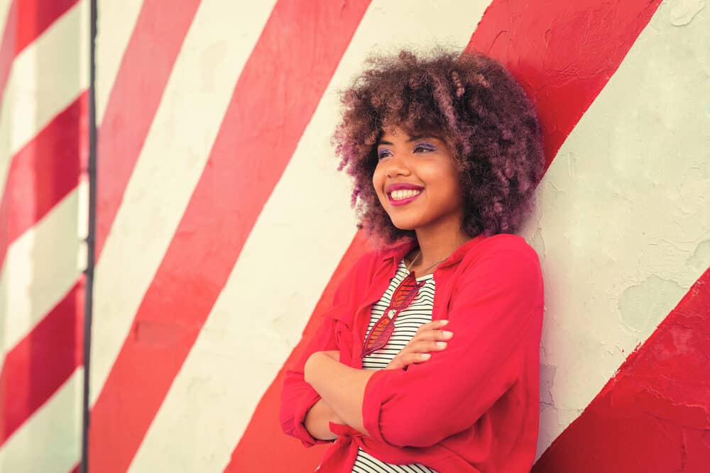 Positive young black woman with curly healthy hair dyed multiple colors soaking up the sun's UV rays outdoors.