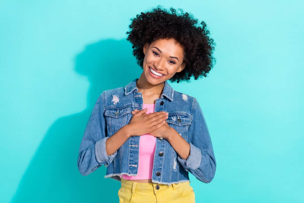 A woman with bleached hair covering her heart with her hands wearing a cut-off blue jean jacket.