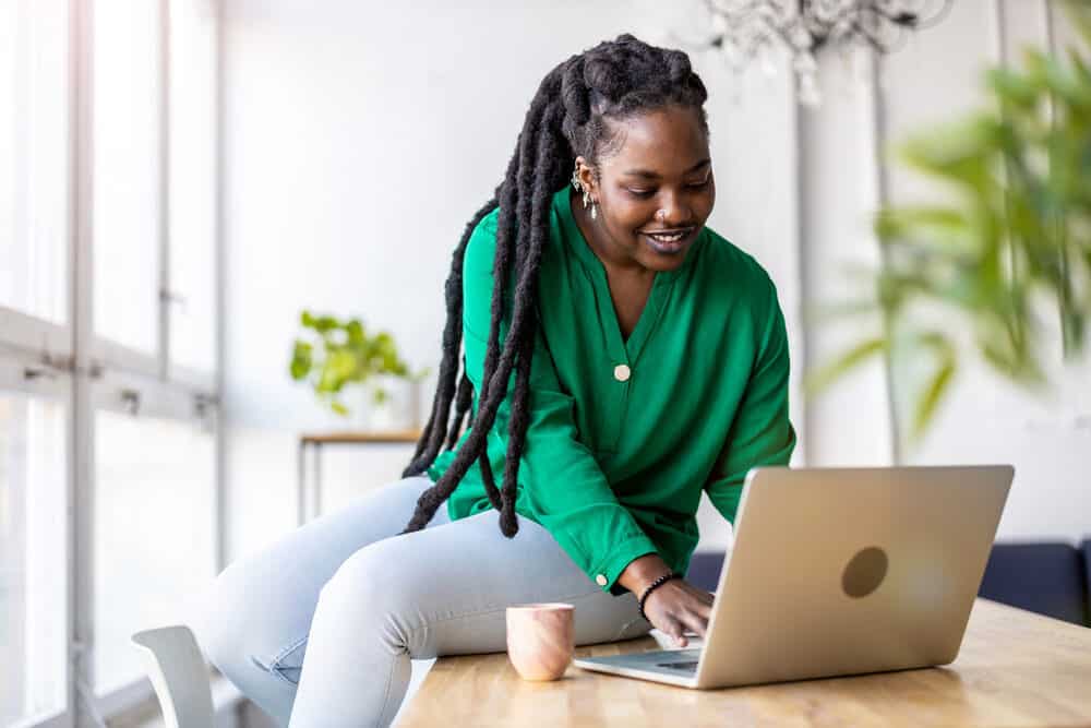 Black female with thinner dreads researching hair loss and healthy roots on her laptop in the kitchen.