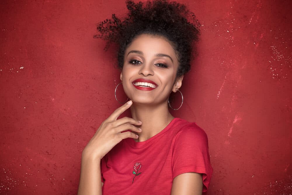 Female with coily hair wearing a wash and go hairdo with red lipstick, hoop earrings, and pink eyeshadow.