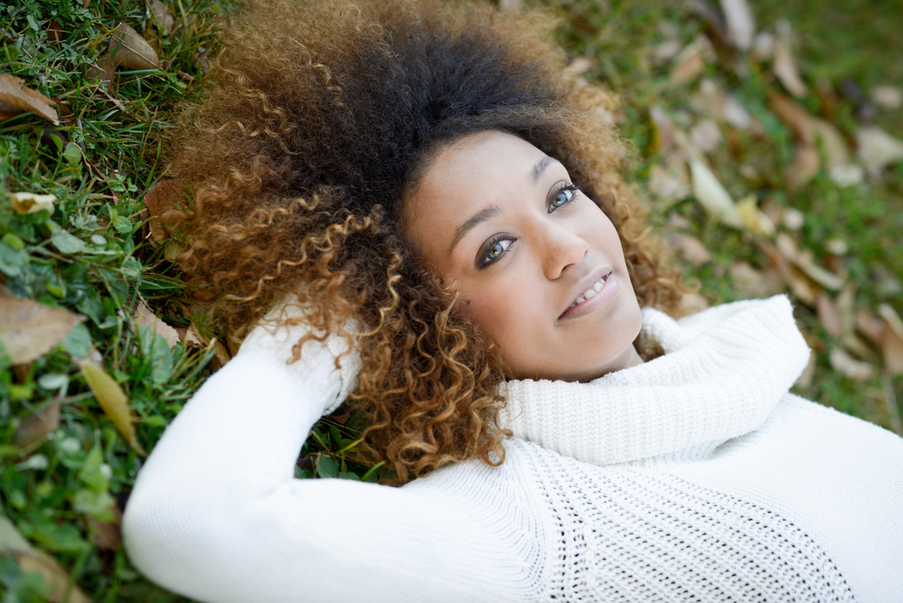 An African American woman with naturally dark hair suffering from hot roots and relaxing on the ground outside.