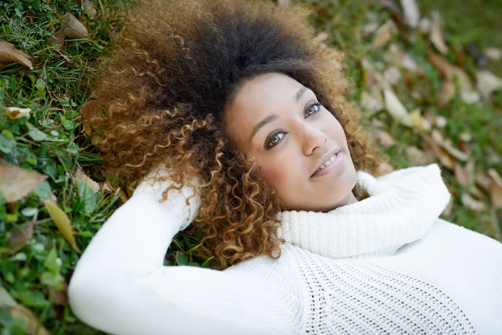 How To Fix Hot Roots at Home With Toner, Box Dye, and More 1 An African American woman with naturally dark hair suffering from hot roots and relaxing on the ground outside.
