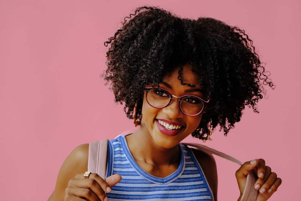 Black lady with thick hair wearing pink and black glasses, red lipstick, and beige backpack.