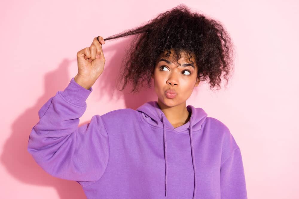Black girl with naturally curly hair suffering from hair fall holding a loose hair thinning from sodium lauryl sulfate.