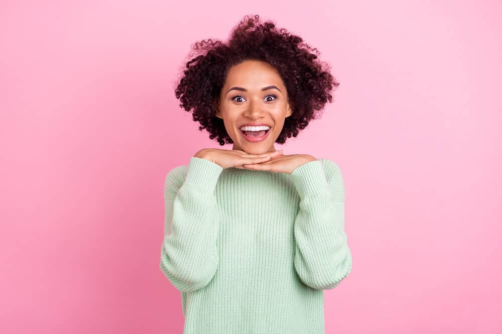 Funny black girl with 3B curly hair holding her hands under her chin wearing a green sweater.