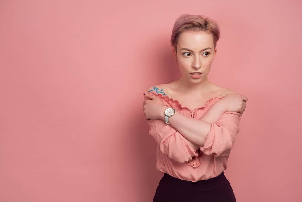 A pretty female with pink hair and a tattoo on her right shoulder wears a blouse and a silver watch.