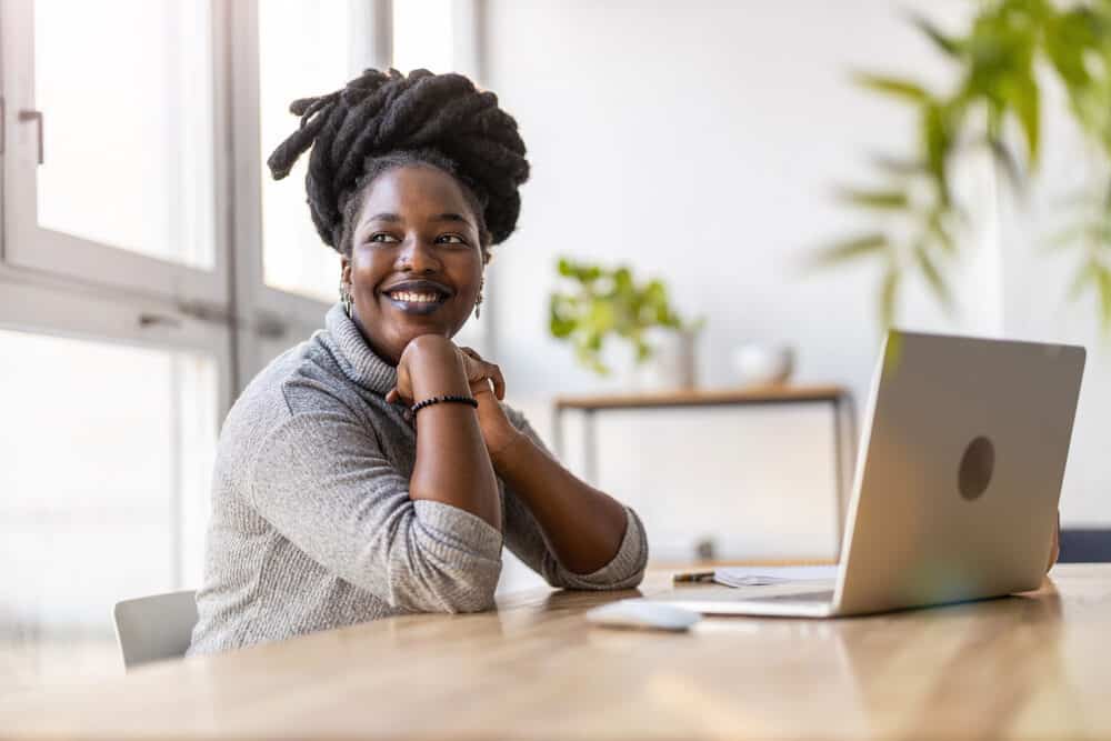 Black lady with a large loc size wearing an updo style using an Apple Macbook laptop to research freeform dreads.