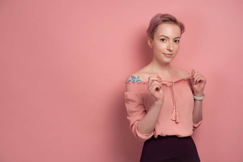 A white lady is pulling the straps on her pink blouse while wearing a subtle smile.