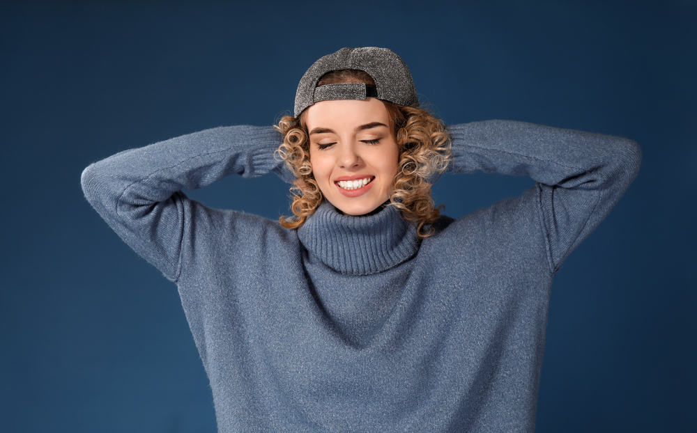 Attractive female with perm dyed hair wearing a blue sweater and baseball cap.