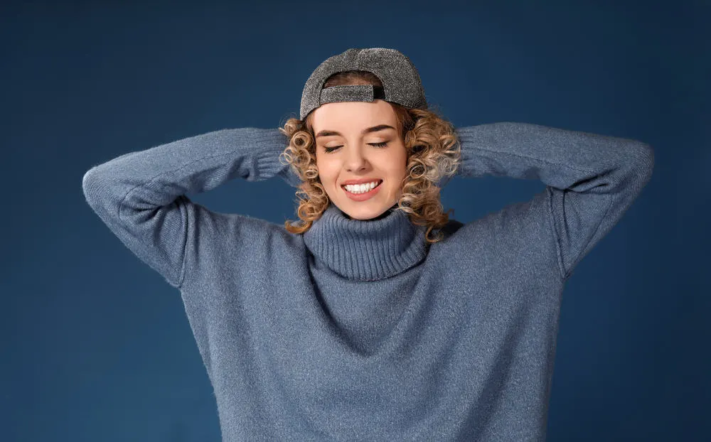Attractive female with perm dyed hair wearing a blue sweater and baseball cap.