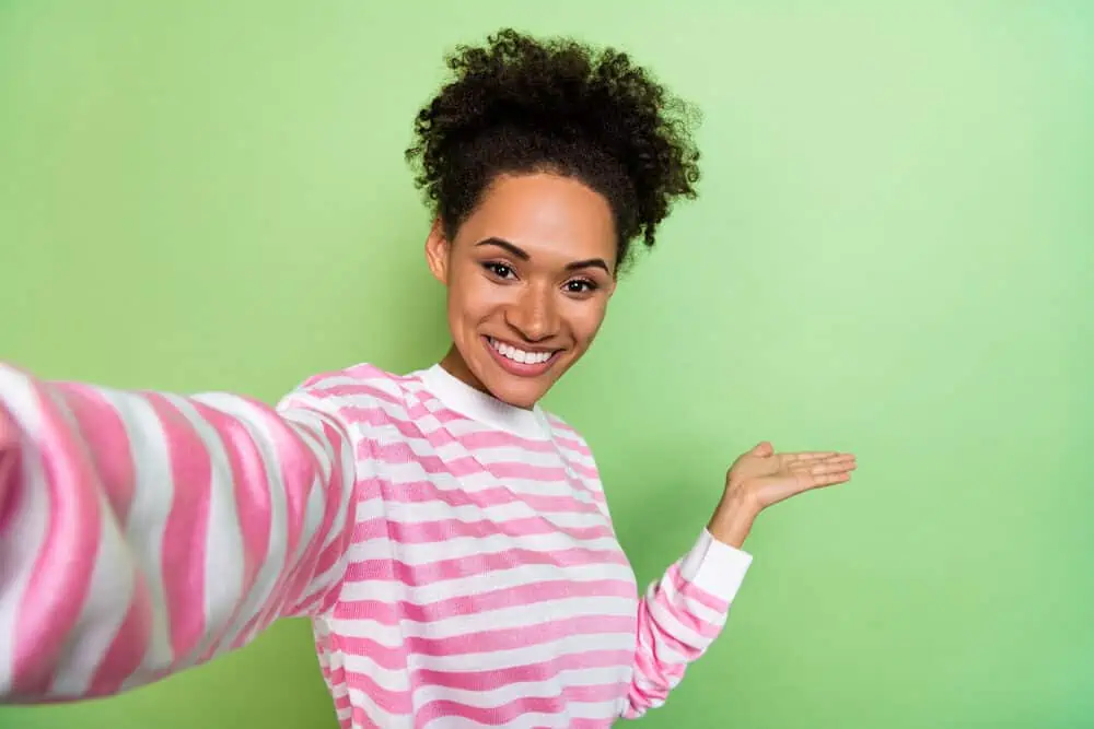 Positive black lady with curly dry hair wearing a pink and white sweater while taking a selfie.