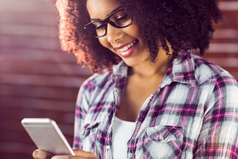 A smiling young black girl researching wig caps, hair toppers, and monofilament wigs on her mobile phone.