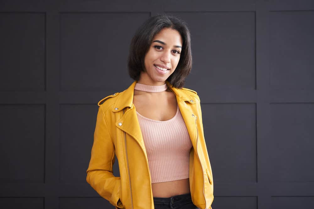 A stylish young girl with thick hair and split ends wearing a yellow leather jacket and pink ribbed shirt.