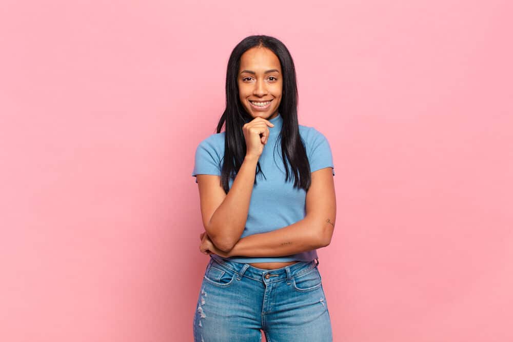 Pretty young black female with jet black straight hair and her hand on chin wearing a blue t-shirt and jeans.