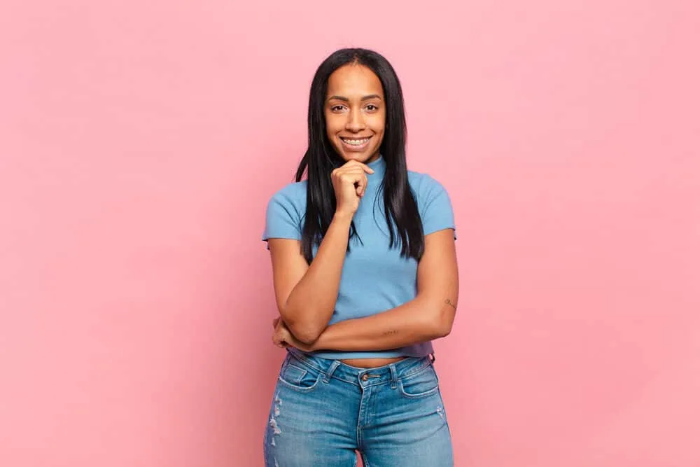 Do Flat Irons Damage Hair? Are Flat Irons Bad for Your Hair? 7 Pretty young black female with jet black straight hair and her hand on chin wearing a blue t-shirt and jeans.