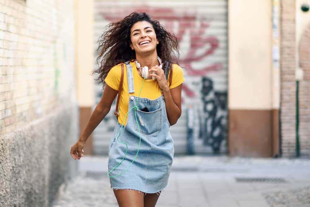 Pretty young African American female with a wavy hair type wearing a blue jean dress and yellow t-shirt.