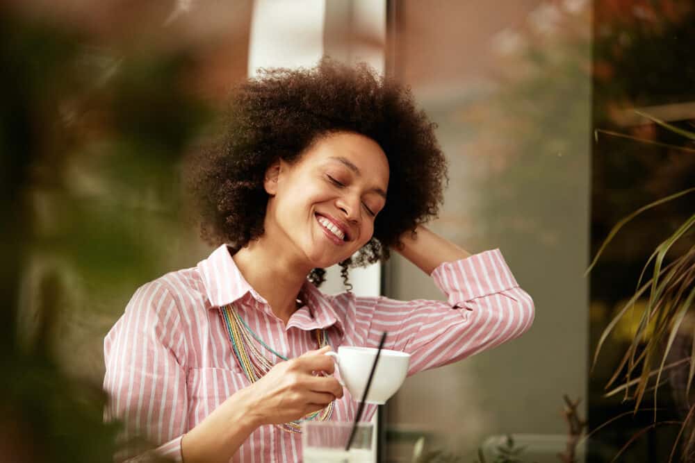 Pretty mixed-race female with dark skin drinking a cup of coffee and wearing a pink and white casual outfit.