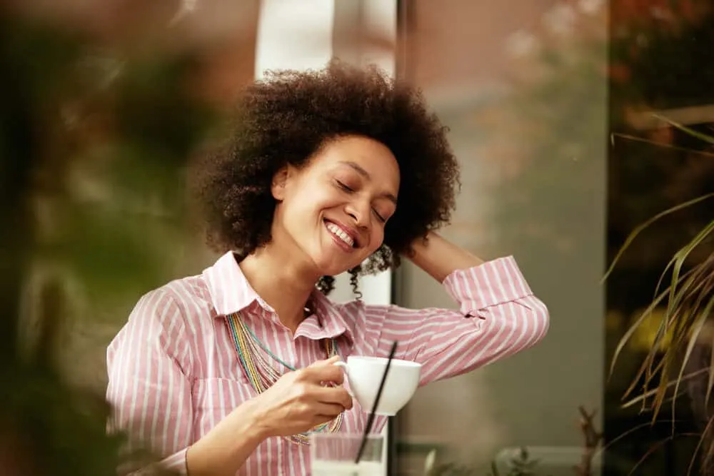 The 11 Best Unnatural Hair Colors for Cool Skin Tones 2 Pretty mixed-race female with dark skin drinking a cup of coffee and wearing a pink and white casual outfit.