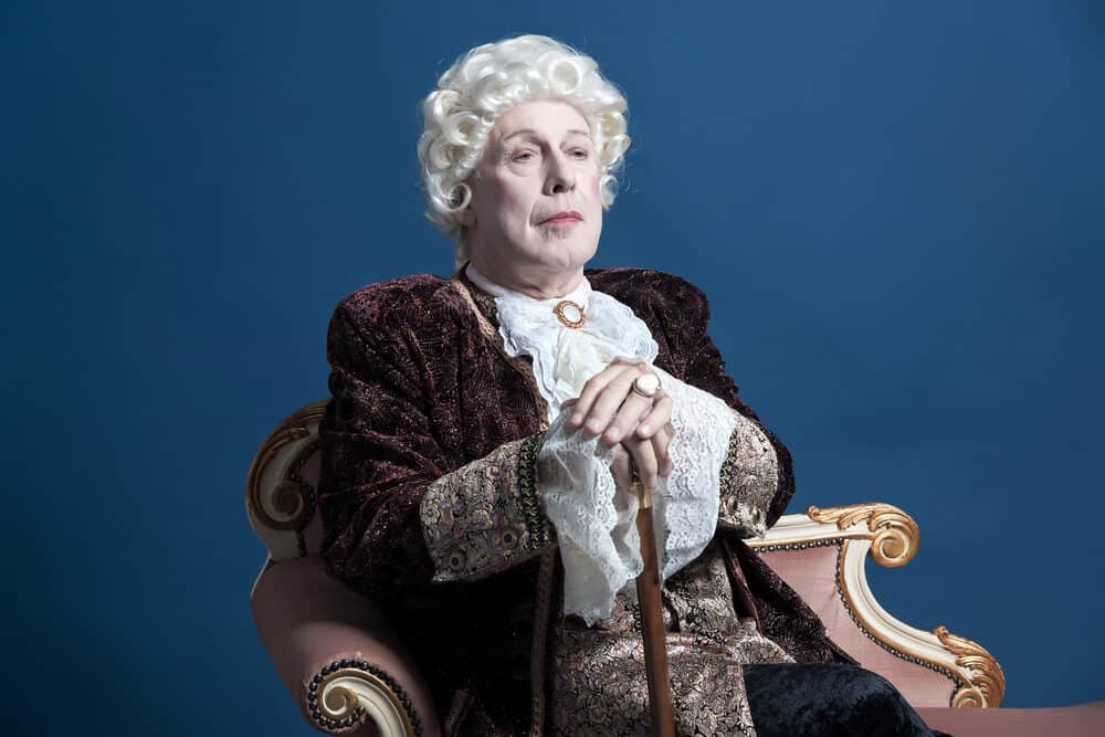 An older Caucasian man dressed as King Louis XIV wearing a human hair wig with white hair covering his own hair.