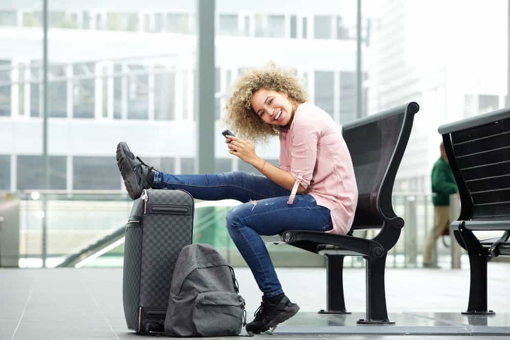 Cute African American female wearing a pink casual shirt, blue jeans, and black sneakers as she waits at the airport.