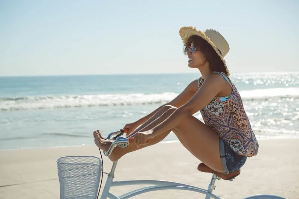 Young black woman with wavy hair wearing casual summer clothes and a straw hat while riding a bike at the beach. 