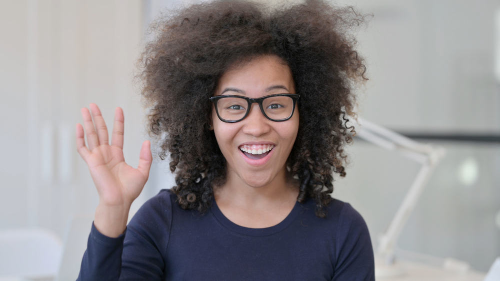 A cute light-skinned black girl with thicker hair has a huge smile while waving with her right hand.