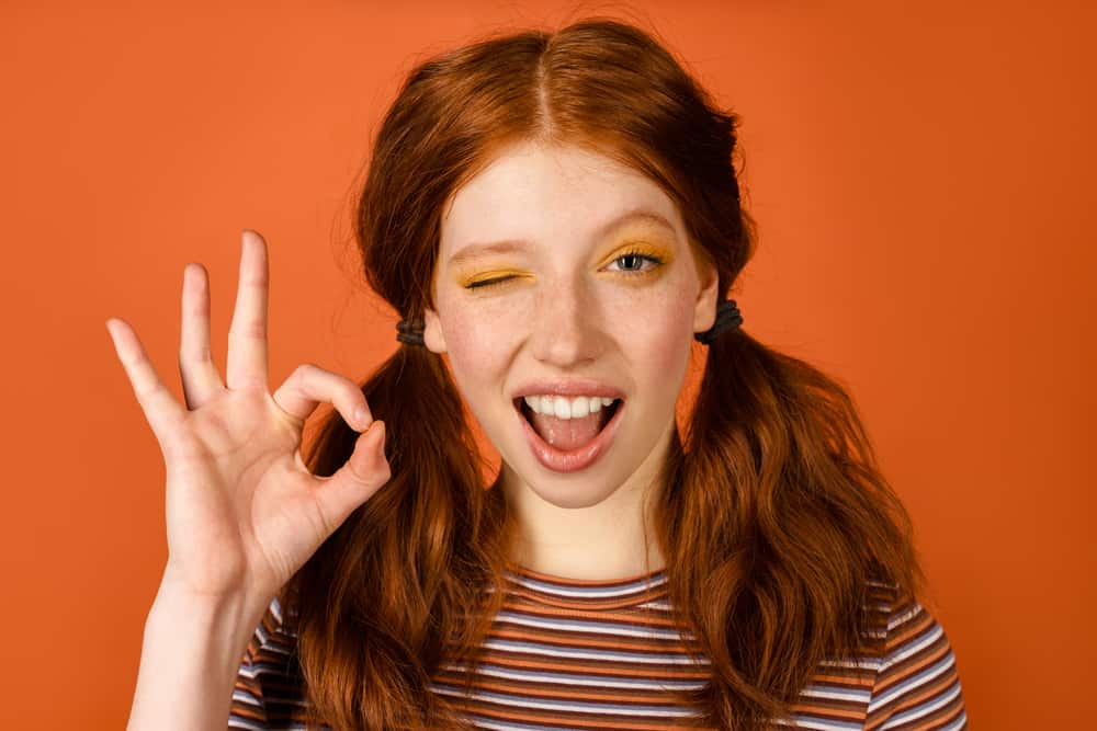 A white girl with pigtails wearing her hair orange and pink lipstick while winking.