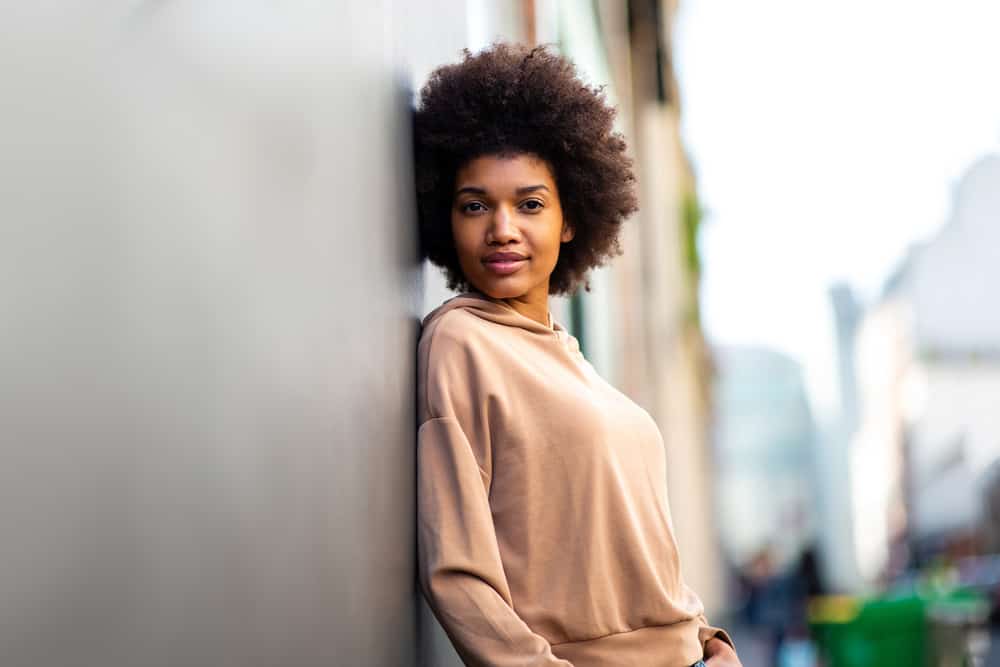 A black female leaning against an outdoor wall wearing her curly long hair in an afro hairstyle.