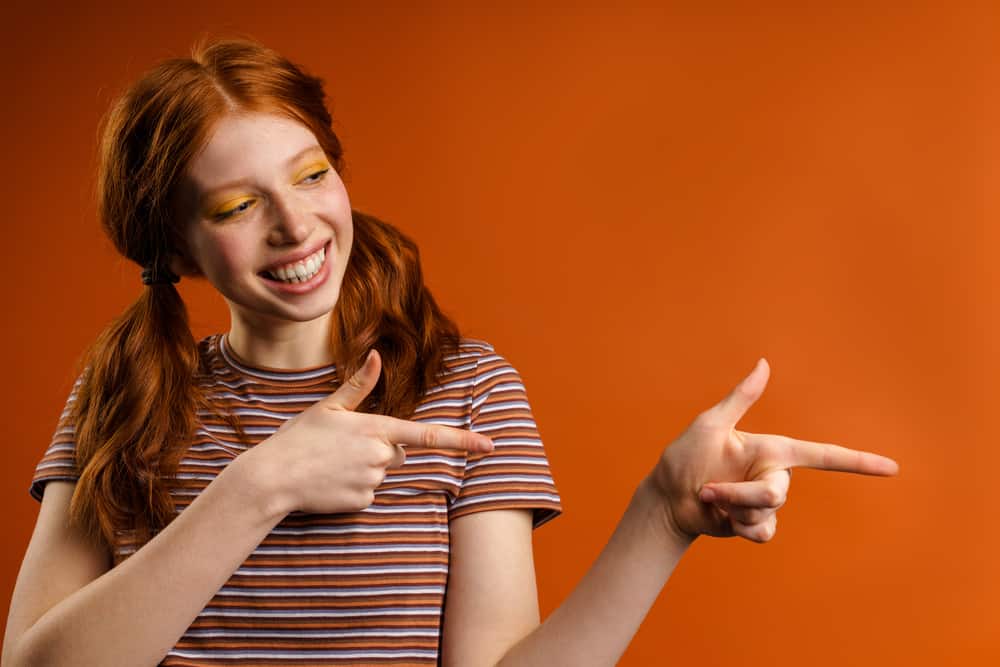 A young woman with bright, orange hair pointing fingers at a friend in the background.